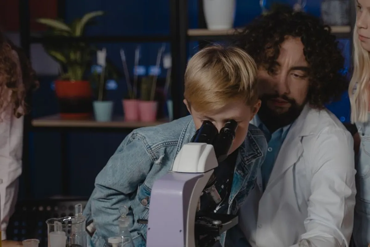 A student looks through a microscope while a teacher guides him, with flasks on the bench beside them