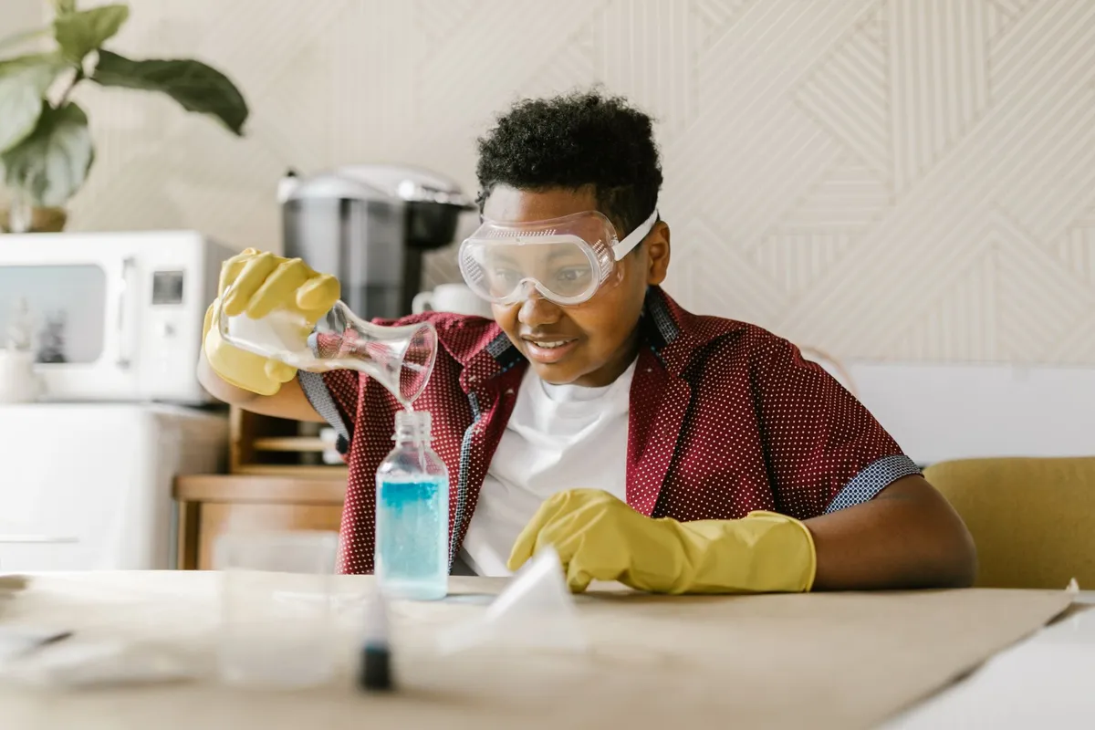 A boy in safety goggles and rubber gloves pours liquid into a bottle at an ordinary table at home