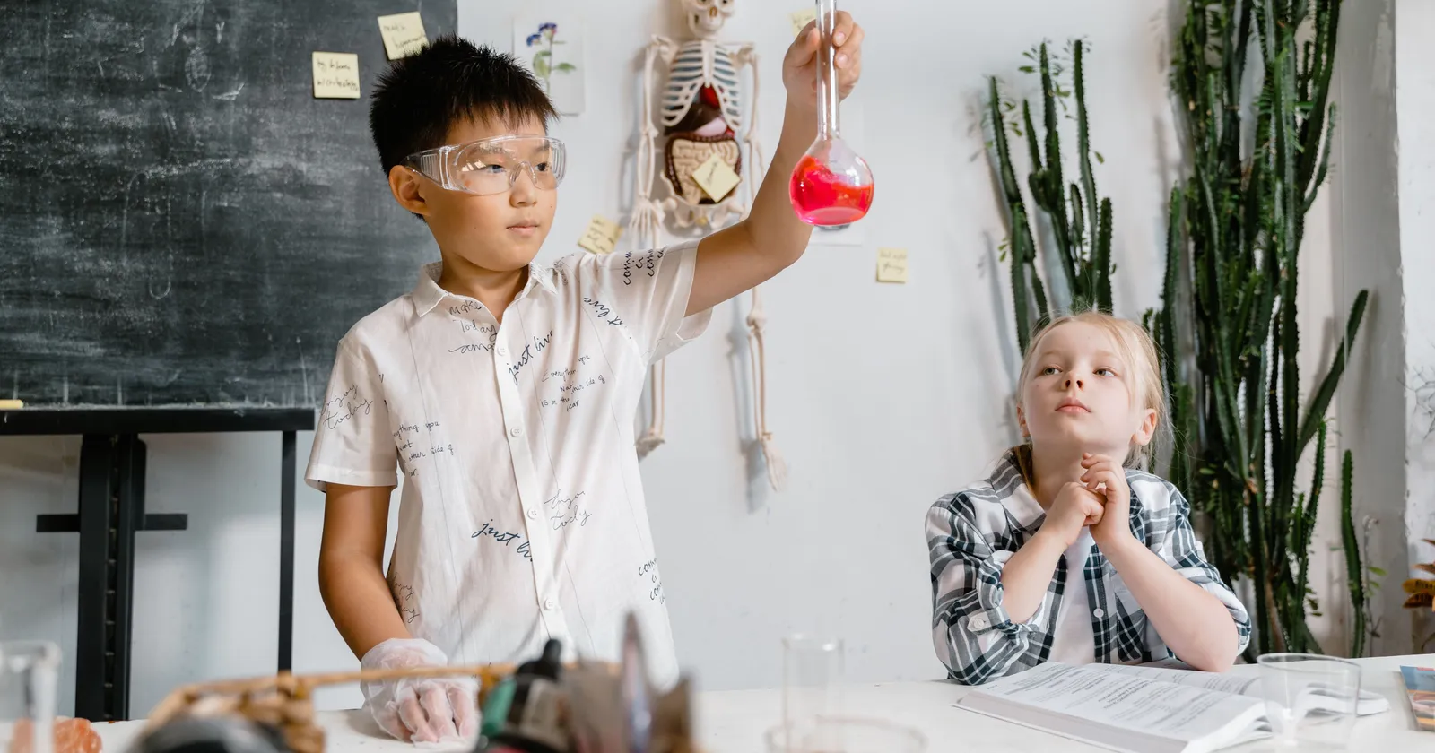 A student in safety goggles holding up a flask of red liquid during a hands-on science experiment, with a classmate watching from the next table